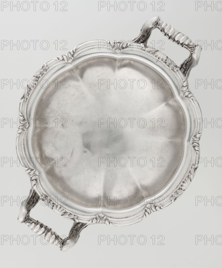 Punch vase, made by Paul Storr 1771–1844 and Storr & Mortimer London; goldsmith's firm, fl. 1822–1838. Spherical, flattened, eight-part burled and ribbed punch vase, supported on four volute leafy feet. Two handles decorated with acanthus leaves and stylized shells. Upper edge turned up and decorated with relief ornament of leaves and shells. Belly engraved with a coat of arms in a decorative cartouche. Materials/technique: burling; silver implied by goldsmith. Date: 1831/1832. Place of origin: London, England, Great Britain. Dimensions: height 15.5 cm; width 36.2 cm; depth 27.8 cm; weight 2490 g., A round metal tray with an ornate scalloped rim and two decorative handles is shown flat against a light background; the tray and handles are metallic silver with areas of subtle darker gray patina, the rim features carved leaflike embellishments and the interior has a softly brushed, slightly mottled silver surface with gentle radial indentations, while the handles have ribbed and textured ...