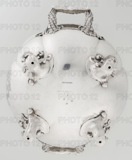 Punch vase, maker: Paul Storr 1771–1844; firm: Storr & Mortimer London; goldsmiths, fl. 1822–1838. English coat of arms engraved on belly in a decorative cartouche. Burling; spherical, flattened, eight-part, burled and ribbed punch vase supported on four volute leafy feet. Two handles decorated with acanthus leaves and stylized shells. Upper edge turned up and decorated with relief ornament of leaves and shells. Materials/technique: silver punch vessel. Date: 1831/1832. Place of origin: London, England, Great Britain. Dimensions: height 15.5 cm; width 36.2 cm; depth 27.8 cm; weight 2490 g., A polished metallic object with a reflective silver surface fills the image, showing curved bulbous forms and ornate sculpted mounts; a pair of decorative handles with textured, rope-like detailing are visible at opposite edges, and four sculpted protrusions with curled ornamental elements extend outward, each ending in a hollow opening; the surface shows small areas of darker reflection and subtle ...