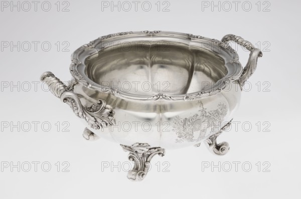 Punch vase, maker: Paul Storr 1771–1844; firm: Storr & Mortimer London; goldsmiths, fl. 1822–1838. Spherical, flattened, eight-part burled and ribbed punch vase supported on four volute leafy feet. Two handles decorated with acanthus leaves and stylized shells. Upper edge turned up and decorated with a relief ornament of leaves and shells. Belly with an engraved coat of arms in a decorative cartouche. Materials/technique: burling. Subjects/ornament: acanthus, shells, volutes, coats of arms. Date: 1831/1832. Place of origin: London, England, Great Britain. Function: punch vessel. Dimensions: height 15.5 cm; width 36.2 cm; depth 27.8 cm; weight 2490 g., A decorative metal bowl with an ornate rim and curved sides sits on a smooth light background, showing a reflective silver surface with polished highlights and soft shadows; it has a pair of elaborately carved handles with scroll and leaf motifs and rests on ornate curled feet with similar decorative detailing, and the interior reflects t...