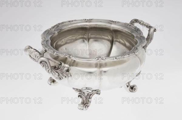 Punch vase by Paul Storr 1771–1844 and Storr & Mortimer London; goldsmiths; fl. 1822–1838. Spherical, flattened, eight-part burled and ribbed punch vase, burling and ribbed surface, supported on four volute leafy feet. Two handles decorated with acanthus leaves and stylized shells. Upper edge turned up and decorated with a relief ornament of leaves and shells. Belly engraved with a coat of arms in a decorative cartouche. Materials/technique: silver goldsmithing. Motifs: acanthus, shells, volutes. Date: 1831/1832. Place of origin: London, England, Great Britain. Function: punch vessel. Dimensions: height 15.5 cm; width 36.2 cm; depth 27.8 cm; weight 2490 g., A polished silver bowl with an open top rests on ornate feet and has decorative handles on opposite sides; the bowl's rim and body feature embossed, curving floral and leaf motifs, and the interior shows reflective silver surfaces with soft highlights and shadows. The visible colors are silver and shades of gray against a light gray...
