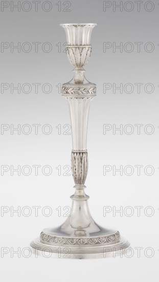 Single-candle candlestick by John Schofield fl. 1776–1796. Classicism/Georgian/George III style. Silver candlestick supported on a circular, stepped-profile foot, decorated with a band of stylised acanthus tendrils with flowers; faceted stem, enclosed at the bottom by lanceolate leaves, widening towards the top and terminating in a ring knurled with oak leaves; vase sleeve with lanceolate leaves at the bottom, faceted above with a slightly rolled-up edge. Dated 1789/1790. England, London. Function: lighting. Height 29.8 cm; diameter 13.8 cm; weight approx. 680 g., A single tall metallic object with a reflective silver surface stands against a light gray background, featuring a flared top with vertical ridges, a rounded midsection with carved leaf-like patterns, a long tapered column with fluted vertical grooves, a narrower ribbed section with elongated leaf motifs, and a broad circular base decorated with a raised band of floral or vine ornamentation; the visible colors are silver and ...