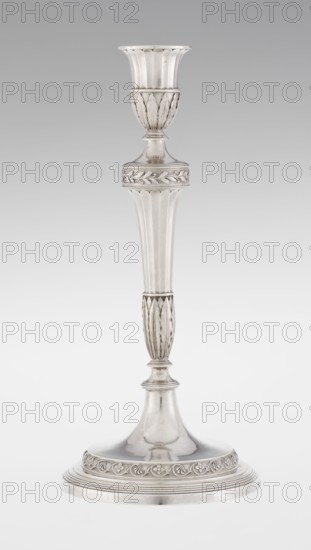 Single-candle candlestick by John Schofield fl. 1776–1796. Classicism/Georgian style with acanthus ornament and oak leaves. The candlestick is supported on a circular, stepped-profile foot decorated with a band of stylised acanthus tendrils with flowers; faceted stem enclosed at the bottom by lanceolate leaves, widening towards the top and terminating in a ring knurled with oak leaves. Vase sleeve with lanceolate leaves at the bottom, faceted above with a slightly rolled-up edge. Dated 1789/1790, London, England. Function: lighting. Height 29.8 cm; diameter 13.8 cm; weight approx. 680 g., A tall metallic candlestick with a reflective silver surface, featuring a flared cup at the top with vertical fluting, a rounded section with embossed leaf-like motifs, a long tapered column with subtle vertical ridges, and a decorative band with raised leaf patterns near the middle; the stem narrows into a smaller fluted section above a rounded joint and rests on a wide, domed base that has concentri...