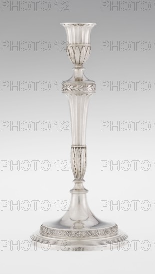 Single-candle candlestick by John Schofield fl. 1776–1796. Classicism/Georgian style with acanthus ornament and oak leaves. Candlestick supported on a circular, stepped-profile foot decorated with a band of stylised acanthus tendrils with flowers; faceted stem enclosed at the bottom by lanceolate leaves, widening towards the top and terminating in a ring knurled with oak leaves; vase sleeve with lanceolate leaves at the bottom, faceted above with a slightly rolled-up edge. Dated 1789/1790. England, London. Function: lighting. Height 29.8 cm, diameter 13.8 cm, weight approx. 680 g., A tall, slender metallic candlestick with a flared cup-shaped top and a narrow stem, shown against a plain light gray background. The candlestick has decorative fluted and leaf-like carved details along the cup, upper shaft, and a lower section of the stem, and a rounded stepped base with a carved band of repeating floral or vine motifs. The surface appears metallic with a polished finish showing reflective ...
