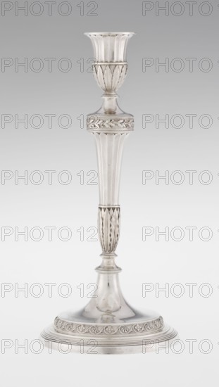 Single-candle candlestick by John Schofield fl. 1776–1796. Classicism/Georgian style with acanthus ornament and oak leaves. The candlestick is supported on a circular, stepped-profile foot decorated with a band of stylised acanthus tendrils with flowers; faceted stem enclosed at the bottom by lanceolate leaves, widening toward the top and terminating in a ring knurled with oak leaves. Vase sleeve with lanceolate leaves at the bottom, faceted above with a slightly rolled-up edge. Dated 1789/1790, London, England. Lighting object. Height 29.8 cm; diameter 13.8 cm; weight approx. 680 g., A tall, slender metallic candlestick with a flared top cup, a narrow stem that widens into a rounded base, and ornate carved banding and fluted details along the upper and lower sections; the surface appears smooth and reflective with a few darker patina areas, and the visible colors are silver, gray, and subtle off-white tones.