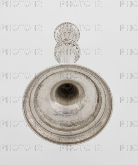 Single-candle candlestick by John Schofield fl. 1776–1796. Classicism/Georgian/George III style; 1789/1790, London, England. Candlestick supported on a circular stepped-profile foot decorated with a band of stylised acanthus tendrils with flowers. Faceted stem, enclosed at the bottom by lanceolate leaves, widening towards the top and terminating in a ring knurled with oak leaves. Vase sleeve with lanceolate leaves at the bottom, faceted above with a slightly rolled-up edge. Materials: metal goldsmith work. Function: lighting. Height 29.8 cm; diameter 13.8 cm; weight approx. 680 g., A close-up view of a metallic object photographed from below showing a flared circular base with concentric ridges and a central hollow, connected to a vertically oriented decorative stem with scalloped, fluted detailing; the visible colors are silver, gray, and subtle dark tarnish tones against a light gray background.