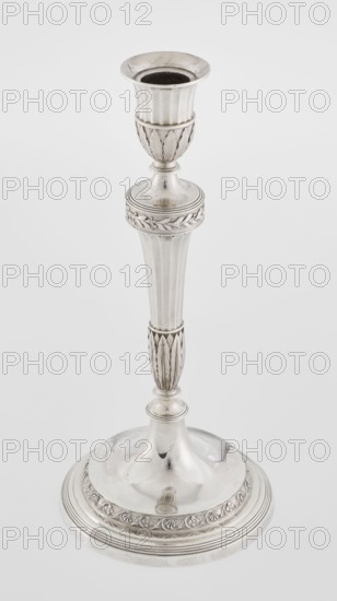 Single-candle candlestick by John Schofield fl. 1776–1796. Classicism/Georgian/George III style. Silver candlestick supported on a circular, stepped-profile foot, decorated with a band of stylised acanthus tendrils with flowers; faceted stem enclosed at the bottom by lanceolate leaves, widening towards the top and terminating in a ring knurled with oak leaves; vase sleeve with lanceolate leaves at the bottom, faceted above with a slightly rolled-up edge. Dated 1789/1790. England, London. Function: lighting. Height 29.8 cm; diameter 13.8 cm; weight approx. 680 g., A tall metallic candlestick with a reflective silver surface stands against a plain light gray background, showing a flared cup at the top, a ribbed upper section, a decorative band with leaf-like motifs, a tapered fluted column, a narrower carved midsection with vertical leaf patterns, and a wide round base with concentric ridges and an ornate frieze; visible colors include silver, gray, and subtle darker tonal reflections.