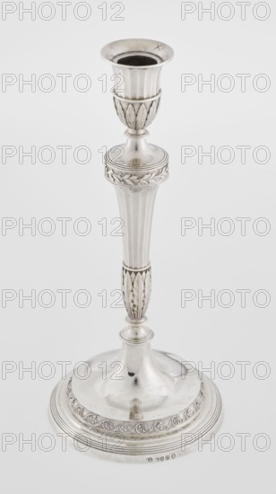 Single-candle candlestick by John Schofield fl. 1776–1796. Classicism/Georgian/George III style. Silver candlestick supported on a circular stepped-profile foot, decorated with a band of stylised acanthus tendrils with flowers; faceted stem enclosed at the bottom by lanceolate leaves, widening toward the top and terminating in a ring knurled with oak leaves; vase sleeve with lanceolate leaves at the bottom, faceted above with a slightly rolled-up edge. Inscribed/dated 1789/1790. England, London. Function: lighting. Height 29.8 cm; diameter 13.8 cm; weight approx. 680 g., A single tall metallic candlestick with a polished silver surface, showing ornamental fluted and leaf-like carved sections along its stem and base; the top has a flared cup with a dark interior, the middle features a rounded knop with a decorative band, and the wide circular base has a raised domed center and a patterned border. The visible colors are silver, metallic gray, and dark gray inside the cup.