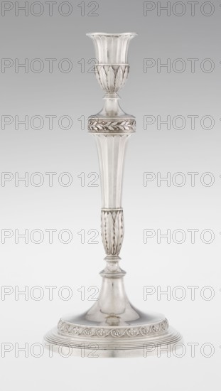 Single-candle candlestick by John Schofield fl. 1776–1796. Classicism/Georgian/George III style. Single-candle candlestick supported on a circular profiled stepped foot, decorated with a knurled band of stylised acanthus twig with flowers on a seeded background; faceted stem enclosed at the bottom by lanceolate leaves, widening towards the top and terminating in a ring decorated with knurled oak leaves; vase sleeve with lanceolate leaves at the bottom, faceted above with a slightly rolled-up edge. Dated 1789/1790. London, England. Material: metal — lighting object. Height 29.8 cm; width 13.8 cm; weight approx. 680 g., A tall, slender metal candlestick with a flared cup at the top, a narrow stem with decorative fluted and leaf-like patterns, and a round stepped base featuring a band of raised ornamental motifs; the object appears smooth and reflective with visible highlights and shadows, and the colors visible are shades of silver, gray, and white.
