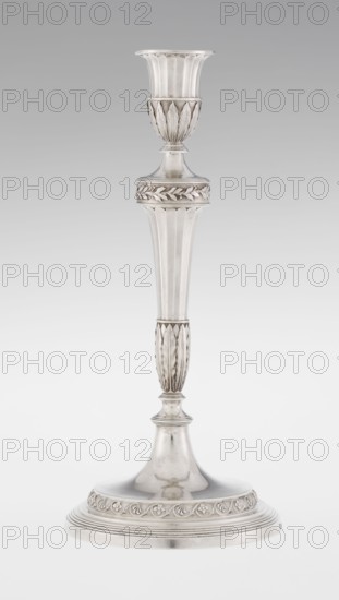 Single-candle candlestick by John Schofield fl. 1776–1796. Classicism/Georgian, George III style. Silver candlestick supported on a circular, profiled, stepped foot, decorated with a knurled band of stylised acanthus twig with flowers on a seeded background; faceted stem enclosed at the bottom by lanceolate leaves, widening towards the top and terminating in a ring decorated with knurled oak leaves; vase sleeve with lanceolate leaves at the bottom, faceted above with a slightly rolled-up edge. Dated 1789/1790, London, England. Function: lighting. Height 29.8 cm; width 13.8 cm; weight approx. 680 g., A tall metallic candlestick stands against a plain light gray background, showing a silvery finish with reflective surfaces and areas of subtle patina; the top has a flared cup with vertical ridges and a decorative band beneath featuring raised leaf-like motifs, followed by a tapered column with fluted vertical ridges and a midsection with repeated elongated leaf patterns, a slender neck an...