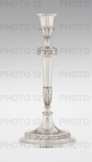 Single-candle candlestick by John Schofield fl. 1776–1796. Georgian/George III classicism style; silver. Supported on a circular profiled stepped foot, decorated with a knurled band of stylized acanthus twig with flowers on a seeded background; faceted stem enclosed at the bottom by lanceolate leaves, widening toward the top and terminating in a ring decorated with knurled oak leaves; vase sleeve with lanceolate leaves at the bottom, faceted above with a slightly rolled-up edge. Inscribed/dated 1789/1790. Origin: London, England. Function: lighting. Height 29.8 cm; width 13.8 cm; weight approx. 680 g., The image shows a tall, slender candlestick with a metallic silver surface standing against a light gray to white gradient background. The candlestick has a flared top cup with vertical ridges and a decorative band beneath it featuring leaf-like reliefs, a tapered column with fluted vertical grooves, and a rounded base section with a raised decorative band of floral or foliate motifs. Th...