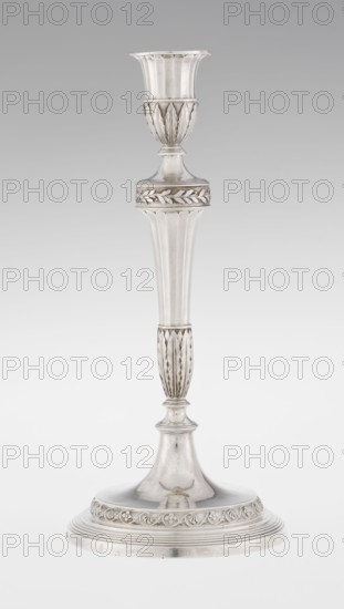 Single-candle candlestick, John Schofield fl. 1776–1796, goldsmith. Classicism/Georgian/George III style; acanthus and oak-leaf ornament. Candlestick supported on a circular profiled stepped foot, decorated with a knurled band of stylised acanthus twig with flowers on a seeded background; faceted stem enclosed at the bottom by lanceolate leaves, widening toward the top and terminating in a ring decorated with knurled oak leaves; vase sleeve with lanceolate leaves at the bottom, faceted above with a slightly rolled edge. Dated 1789/1790. England, London. Lighting. Height 29.8 cm; width 13.8 cm; weight approx. 680 g., A tall metallic candlestick with a reflective silver surface featuring a flared cup at the top and a rounded base, decorated with carved leaf-like and scalloped motifs; a few ribbed vertical sections separate the ornate banding, and the overall color palette shows shades of silver and gray against a pale gray background.