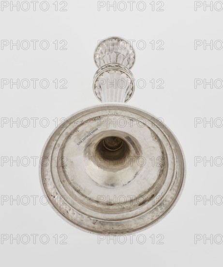 Single-candle candlestick by John Schofield fl. 1776–1796. Georgian/George III classicism style, 1789/1790, London, England. Silver candlestick supported on a circular, profiled, stepped foot, decorated with a knurled band of stylised acanthus twig with flowers on a seeded background; faceted stem enclosed at the bottom by lanceolate leaves, widening toward the top and terminating in a ring decorated with knurled oak leaves; vase sleeve with lanceolate leaves at the bottom, faceted above with a slightly rolled-up edge. Ornament: acanthus, oak leaves. Function: lighting. Height 29.8 cm; width 13.8 cm; weight approx. 680 g., A close-up view of a metallic object photographed from below showing a flared circular base with concentric ridges and a central hollow, connected to a decorative stem with scalloped and fluted patterns; the object appears in shades of silver and gray against a plain white background.