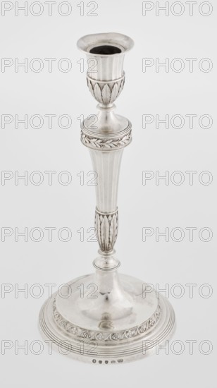 Single-candle candlestick by John Schofield fl. 1776–1796. Classicism/Georgian, George III style. Silver candlestick supported on a circular profiled stepped foot, decorated with a knurled band of stylised acanthus twig with flowers on a seeded background; faceted stem enclosed at the bottom by lanceolate leaves, widening towards the top and terminating in a ring decorated with knurled oak leaves; vase sleeve with lanceolate leaves at the bottom, faceted above with a slightly rolled-up edge. Dated 1789/1790, London, England. Function: lighting. Height 29.8 cm; width 13.8 cm; weight approx. 680 g., A silver-colored metal candlestick stands upright against a plain light gray background; it has a flared cup at the top with a darker interior, a decorative midsection with leaf-like vertical ridges and a rounded collar with embossed leaf motifs, a tapered fluted stem with a narrower carved section repeating the leaf motif, and a broad circular base with concentric ridges and an embossed deco...