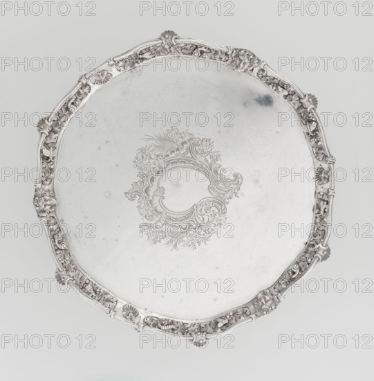 Leg tray. Maker: Robert Harper fl. 1853–1869; firm: Harper & Son London; goldsmith's firm; fl. 1869–1889. Materials/technique: circular patera vessel. Decoration/style: Neo-Rococo; engraved and relief ornament including rocailles, rocaille, rooster combs, acanthus volutes, shells, volutes, floral sprigs, vine leaves and grapes, high-relief insects butterflies, moths, midges, snails, a caterpillar, and four Dionysus masks. Description: supported on three volute legs terminating in vine leaves at the base; mirror with blind reserve richly decorated with engraved neo-Rococo ornament; slightly raised openwork collar with relief vine leaves and grapes and high-relief insects and mollusks; collar edge profiled with half-cylinder decorated with shells and rooster comb. Date: 1860–1861. Place: London, England, Great Britain. Function: vessel. Dimensions: height 3.9 cm; diameter 32.5 cm; weight 895 g., A round metallic tray with a scalloped, ornate rim featuring a decorative floral and shell mo...