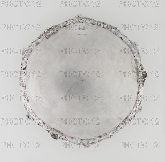 Leg tray by Robert Harper fl. 1853–1869, maker; Harper & Son London, goldsmith's firm, fl. 1869–1889. Circular patera supported on three volute legs terminating in vine leaves at the base. The mirror features a blind reserve richly decorated with engraved Neo-Rococo ornament in the form of rocailles, rooster combs, acanthus volutes, and floral sprigs. The slightly raised, openwork collar features relief decoration of vine leaves and grapes, with four Dionysus masks at the feet, and high-relief insects butterflies, moths, midges, snails, and a caterpillar. The collar edge is profiled with a half-cylinder decorated with shells and a rooster comb. Subject/iconography: head of Dionysus; motifs: insects, vines, snails, caterpillars, rocaille, shells, volutes, acanthus leaf. Date: 1860–1861. Place: London, England, Great Britain. Medium/object type: vessel leg tray. Dimensions: height 3.9 cm; diameter 32.5 cm; weight 895 g., A round metallic tray viewed from above with a smooth central surfa...