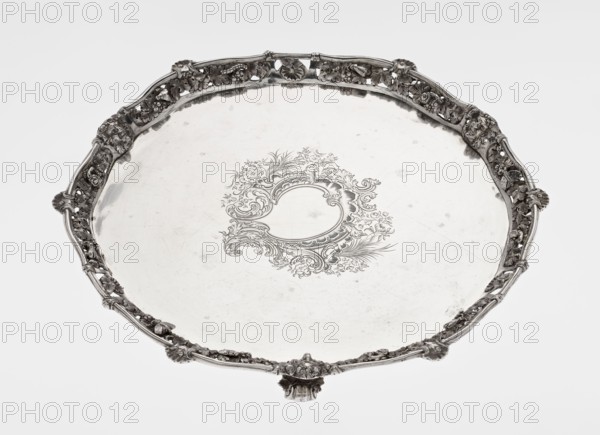 Leg Tray by Robert Harper fl. 1853–1869 and Harper & Son London; goldsmith's firm; fl. 1869–1889. Circular patera supported on three volute legs terminating in vine leaves at the base. The mirror features a blind reserve richly decorated with engraved Neo-Rococo ornament in the form of rocailles, rooster combs, acanthus volutes, and floral sprigs. The slightly raised, openwork collar features relief decoration of vine leaves and grapes, with four Dionysus masks at the feet, and high-relief insects butterflies, moths, midges, snails, and a caterpillar. The collar edge is edged with a profiled half-cylinder decorated with shells and a rooster comb. Inscriptions/themes: insects, vines, snails, caterpillars, head of Dionysus, rocaille, volutes, acanthus leaf. Date: 1860/1861. Place: London, England, Great Britain. Medium/function: vessel. Dimensions: height 3.9 cm; diameter 32.5 cm; weight 895 g., A round decorative tray with an ornate raised rim and sculpted edge, the tray and rim appeari...