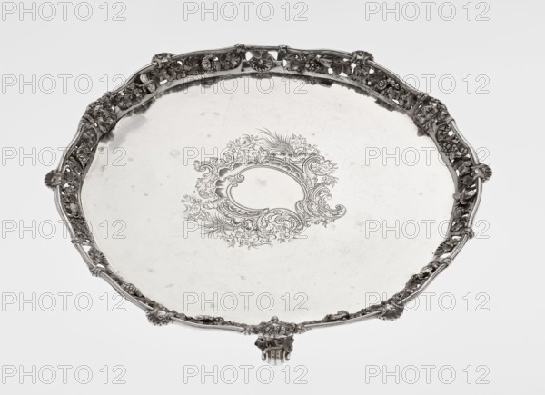 Leg Tray. Maker: Robert Harper fl. 1853–1869; firm: Harper & Son London; goldsmith's firm; fl. 1869–1889. Materials/subject: Circular patera supported on three volute legs terminating in vine leaves at the base. The mirror features a blind reserve richly decorated with engraved Neo-Rococo ornament in the form of rocailles, rooster combs, acanthus volutes, and floral sprigs. The slightly raised, openwork collar features relief decoration of vine leaves and grapes, with four Dionysus masks at the feet, and high-relief insects butterflies, moths, midges, snails, and a caterpillar. The collar edge is edged with a profiled half-cylinder decorated with shells and a rooster comb. Iconography: head of Dionysus. Style: Neo-Rococo rocaille, volutes, acanthus leaf. Date: 1860–1861. Place: London, England, Great Britain. Object type: vessel. Dimensions: height 3.9 cm; diameter 32.5 cm; weight 895 g., A large round metal tray with an ornate raised rim and a decorative engraved motif in the center, ...