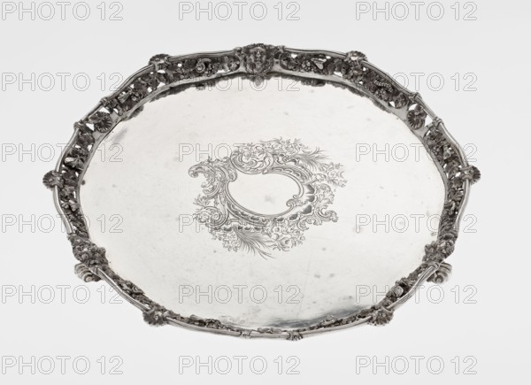 Leg Tray by Robert Harper fl. 1853–1869; Harper & Son London; goldsmith's firm; fl. 1869–1889. Circular patera supported on three volute legs terminating in vine leaves at the base. The mirror features a blind reserve richly decorated with engraved Neo-Rococo ornament in the form of rocailles, rooster combs, acanthus volutes, and floral sprigs. The slightly raised, openwork collar bears relief decoration of vine leaves and grapes, with four Dionysus masks at the feet, and high-relief insects butterflies, moths, midges, snails, and a caterpillar. The collar edge is profiled with a half-cylinder decorated with shells and a rooster comb. Iconography: head of Dionysus. Materials/ornament: insects, vines, snails, caterpillars, rocaille, shells, volutes, acanthus leaf; style: Neo-Rococo. Date: 1860/1861. Place: London, England, Great Britain. Vessel. Height: 3.9 cm; diameter: 32.5 cm; weight: 895 g., A round decorative tray with an ornate raised rim featuring floral and scrollwork motifs and...