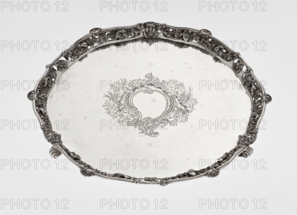 Leg Tray. Maker: Robert Harper fl. 1853–1869; firm: Harper & Son London; goldsmith's firm; fl. 1869–1889. Materials/technique: circular patera supported on three volute legs terminating in vine leaves at the base. The mirror features a blind reserve richly decorated with engraved Neo-Rococo ornament in the form of rocailles, rooster combs, acanthus volutes, and floral sprigs. The slightly raised, openwork collar features relief decoration of vine leaves and grapes, with four Dionysus masks at the feet, and high-relief insects butterflies, moths, midges, snails, and a caterpillar. The collar edge is edged with a profiled half-cylinder decorated with shells and a rooster comb. Iconography: head of Dionysus. Style: Neo-Rococo rocaille, volutes, acanthus leaf. Date: 1860–1861. Place: London, England, Great Britain. Object type: vessel. Dimensions: height 3.9 cm; diameter 32.5 cm; weight 895 g., A large round metal tray with an ornate raised rim that has detailed openwork and sculpted decor...