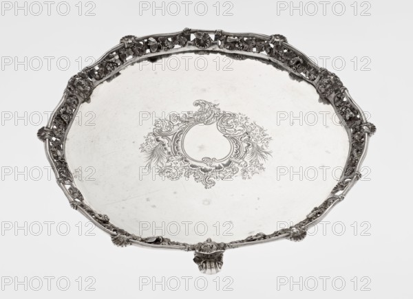 Leg tray by Robert Harper fl. 1853–1869 and Harper & Son London; goldsmith's firm; fl. 1869–1889. Circular patera supported on three volute legs terminating in vine leaves at the base. The mirror features a blind reserve richly decorated with engraved Neo-Rococo ornament in the form of rocailles, rooster combs, acanthus volutes, and floral sprigs. The slightly raised, openwork collar has relief decoration of vine leaves and grapes, with four Dionysus masks at the feet, and high-relief insects butterflies, moths, midges, snails, and a caterpillar. The collar edge is profiled half-cylinder decorated with shells and a rooster comb. Motifs: insects, vines, snails, caterpillars, head of Dionysus; rocaille, volutes, acanthus leaf ornament. Date: 1860/1861. Place: London, England. Medium/function: vessel leg tray. Dimensions: height 3.9 cm; diameter 32.5 cm; weight 895 g., A round decorative metal tray with an ornate raised rim featuring a repeating floral and scroll motif in metallic silver ...