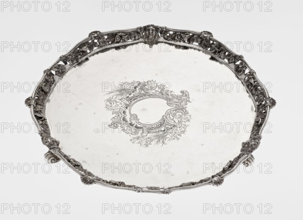 Leg tray by Robert Harper fl. 1853–1869 and Harper & Son London; goldsmith's firm, fl. 1869–1889. Circular patera supported on three volute legs terminating in vine leaves at the base. The mirror features a blind reserve richly decorated with engraved Neo-Rococo ornament in the form of rocailles, rooster combs, acanthus volutes, and floral sprigs. The slightly raised, openwork collar features relief decoration of vine leaves and grapes, with four Dionysus masks at the feet, and high-relief insects butterflies, moths, midges, snails, and a caterpillar. The collar edge is edged with a profiled half-cylinder decorated with shells and a rooster comb. Materials/ornament: insects, vines, snails, caterpillars, head of Dionysus, rocaille, shells, volutes, acanthus leaf. Date: 1860–1861. Place: London, England. Vessel. Height 3.9 cm; diameter 32.5 cm; weight 895 g., A round metallic serving tray with an ornate raised rim featuring openwork floral and scroll motifs and several sculpted shell-lik...