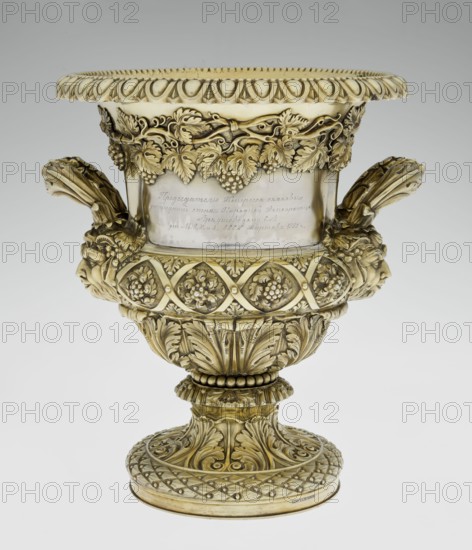 Wine Cooler, Boulton & Plate Co Birmingham; goldsmith and plater firm; fl. 1768–1834 maker; vine ornament; dedications; jockey on horse iconographic; horse racing; classicism style; Georgian style; acanthus leaf ornament; Satyr heads iconographic; cymatium ornament. Classicist on a circular foot decorated with a half-cylinder of laurel and acanthus leaves overlapping a short stem; body enclosed below with large acanthus leaves, above decorated with vines in a diagonal, rhombic braid. Plaque with racers on the front riders; on the back a plaque with a secondary inscription in Cyrillic script is attached the original inscription, probably referring to a prize in horse races, has been erased. Under the upper edge a decorative strip of vines; the rim is edged with a stylized kimation; on the sides massive ears mounted in satyr masks. Date: 1826/1827. Place of manufacture: Birmingham, England, Great Britain. Object type: vessel wine cooler. Dimensions: height 30 cm; width 27 cm; depth 24.5 ...