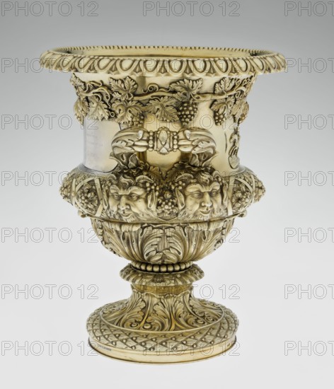 Wine cooler by Boulton & Plate Co. Birmingham; goldsmith and plater firm; fl. 1768–1834. Classicist/Georgian style, 1826/1827. Silver vessel, height 30 cm, width 27 cm, depth 24.5 cm, weight 3725 g. On a circular foot decorated with a half-cylinder of laurel and acanthus leaves overlapping a short stem; body enclosed below with large acanthus leaves, above decorated with vines in a diagonal rhombic braid. Front plaque with horse racers; a secondary plaque with an inscription in Cyrillic script is attached on the back original inscription, probably referring to a prize in horse races, has been erased. Upper edge with a decorative strip of vines; rim edged with a stylized cymatium; sides with massive ears mounted in satyr masks. Ornamentation includes vine, acanthus leaf, cymatium, satyr heads; iconography includes jockey on horse and horse racing., A decorative ornate vessel with a metallic sheen in gold and pale brass tones, featuring a wide flared rim with repeating rounded motifs, a ...