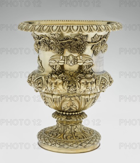 Wine cooler by Boulton & Plate Co. Birmingham; goldsmith and plater firm; fl. 1768–1834. Silver vessel, 1826/1827. Classicism, Georgian style. Circular foot decorated with a half-cylinder of laurel and acanthus leaves overlapping a short stem; body enclosed below with large acanthus leaves, above decorated with diagonal vine in a rhombic braid. Plaque on the front with racers jockey on horse; horse racing; a secondary plaque on the back bears an inscription in Cyrillic script the original inscription, probably referring to a prize in horse races, has been erased. Decorative strip of vines under the upper edge; rim edged with a stylized cymatium/kimation; massive handles mounted in satyr masks. Decorations include vine ornament, acanthus leaf, satyr heads, and classicist motifs. Height 30 cm; width 27 cm; depth 24.5 cm; weight 3725 g., A decorative ornate urn with a metallic golden color overall, featuring a wide flared rim with a repeating scalloped edge and carved teardrop motifs, a b...