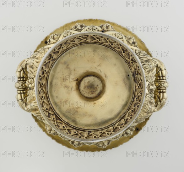 Wine cooler by Boulton & Plate Co Birmingham; goldsmith and plater firm; fl. 1768–1834, 1826/1827. Silver vessel, height 30 cm, width 27 cm, depth 24.5 cm, weight 3725 g. Classicism/Georgian style. Circular foot decorated with a half-cylinder of laurel and overlapping acanthus leaves on a short stem; body enclosed below with large acanthus leaves, above decorated with vines in a diagonal rhombic braid. Plaque with horse racers on the front; a secondary plaque with an inscription in Cyrillic on the back original inscription, probably referring to a prize in horse races, has been erased. Upper edge with a decorative strip of vines; rim edged with a stylized cymatium; massive handles mounted in satyr masks. Ornament: vine, acanthus leaf, cymatium; iconography: jockey on horse, horse racing, satyr heads., A top-down view of an ornate metallic vessel with a central circular depression, surrounded by an intricate raised rim with lattice-like patterns and floral motifs; the piece shows a mix ...