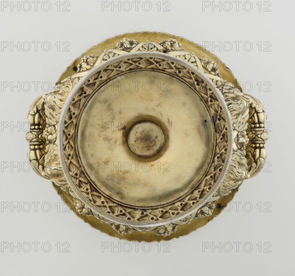 Wine cooler by Boulton & Plate Co. Birmingham; goldsmith and plater firm; fl. 1768–1834. Silver, 1826/1827. Classicism/Georgian style. Circular foot decorated with a half-cylinder of laurel and acanthus leaves overlapping a short stem; body enclosed below with large acanthus leaves, above decorated with vines in a diagonal rhombic braid. Plaque with racers on the front riders; a plaque with a secondary inscription in Cyrillic script is attached on the back the original inscription, probably referring to a prize in horse races, has been erased. Decorative strip of vines under the upper edge; rim edged with a stylized kimation; massive handles mounted in satyr masks. Ornamentation includes vine, acanthus leaf, cymatium, satyr heads; iconography: jockey on horse, horse racing. Vessel. Height 30 cm, width 27 cm, depth 24.5 cm, weight 3725 g., A metallic decorative object viewed from above with a central shallow bowl area in aged gold tones surrounded by an ornate rim with carved patterns, ...