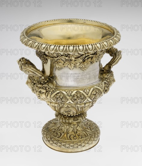 Wine cooler by Boulton & Plate Co Birmingham; goldsmith and plater firm; fl. 1768–1834. Classicist, Georgian style, 1826/1827. Silver vessel, height 30 cm, width 27 cm, depth 24.5 cm, weight 3725 g. Circular foot decorated with a half-cylinder of laurel and acanthus leaves overlapping a short stem; body enclosed below with large acanthus leaves, above decorated with vines in a diagonal rhombic braid. Plaque on the front with horse racers; on the back a plaque with a secondary inscription in Cyrillic script is attached the original inscription, probably referring to a prize in horse races, has been erased. Under the upper edge a decorative strip of vines; rim edged with a stylized cymatium; sides with massive ears mounted in satyr masks. Ornament: vine, acanthus leaf, cymatium; iconography: jockey on horse, horse racing, satyr heads; corporate mottoes and maker’s mark noted., A detailed ornate chalice-like vessel with a wide flared rim and a rounded bowl, featuring elaborate relief deco...