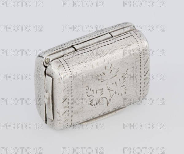Fragrance container vinaigrette, by Thomas Shaw fl. 1822–ca. 1840; 1782–1845. Silver vinaigrette in the form of a bag with an envelope closure and a protruding tongue, decorated on the front with dot-engraved flowers and leaves. Hinged lid opens to reveal an openwork insert decorated with plant scrolls. Dated 1834/1835. Birmingham, England. Toilet accessory. Dimensions: height 2.1 cm, width 1.5 cm, depth 1 cm; weight 10 g., A small rectangular metal box with rounded edges sits angled on a light background, showing a hinged top and a clasp on one side; the surface is a dull silver color with visible scratches and a pattern of incised decorative marks including floral motifs and dotted border lines; there is a small metal hinge and a thin metal strip along one side, and the box casts a faint shadow on the pale surface.