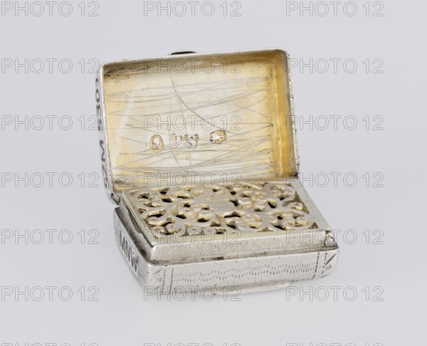 Fragrance container vinaigrette, maker Thomas Shaw fl. 1822–ca. 1840; 1782–1845, goldsmith; scrap-purchase provenance. Vinaigrette in the form of a bag with an envelope closure and a protruding tongue, decorated on the front with dot-engraved flowers and leaves. Hinged lid opening to an interior with an openwork insert decorated with plant scrolls. Dated 1834/1835. England, Birmingham. Toilet accessory. Materials/technique: engraving, openwork. Dimensions: height 2.1 cm, width 1.5 cm, depth 1 cm; weight 10 g., A small open metal box with a hinged lid shown from an angle, the interior of the lid displaying scratches and a worn golden surface, the box interior featuring a decorative filigree insert with ornate cutouts and touches of gold, the exterior of the box appearing silver with engraved patterns and a slightly tarnished, aged finish; the overall colors visible are silver, gold, and pale gray, set against a light gray background.