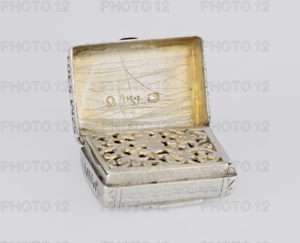 Fragrance container vinaigrette in the form of a bag with an envelope closure and a protruding tongue, decorated on the front with dot-engraved flowers and leaves. Opening box with hinged lid; inside openwork insert decorated with plant scrolls. Maker: Thomas Shaw fl. 1822–ca. 1840; 1782–1845, goldsmith. Date: 1834/1835. Material: scrap metal. Style/decoration: engraving, openwork, plant/flower ornaments; shape: bag. Function: vinaigrette/toilet accessory. Dimensions: height 2.1 cm, width 1.5 cm, depth 1 cm; weight 10 g. Place: Birmingham, England, Great Britain. Provenance: purchase., A small open rectangular metal box is shown against a light gray background, with its hinged lid raised to reveal an interior that has a warm golden tone and visible surface scratches; the inside of the lid has three small raised oval marks. The lower half features a decorative perforated panel with an ornate floral or scrollwork pattern finished in a combination of silvery metal and touches of gold, and...
