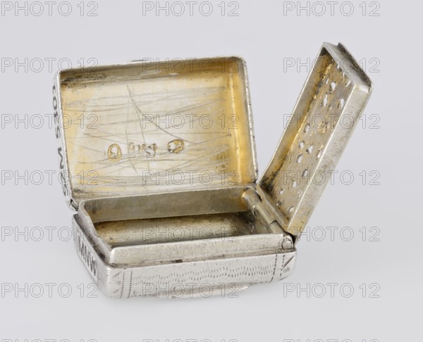 Fragrance container vinaigrette, maker: Thomas Shaw fl. 1822–ca. 1840; 1782–1845, goldsmith. Vinaigrette in the form of a bag with an envelope closure and a protruding tongue, decorated on the front with dot-engraved flowers and leaves. Hinged lid opens to an inner openwork insert decorated with plant scrolls. Materials/technique: engraving, openwork. Date: 1834/1835. Place: Birmingham, England, Great Britain. Function: toilet accessory. Dimensions: height 2.1 cm; width 1.5 cm; depth 1 cm; weight 10 g., A small open metal box with two hinged lids is shown resting on a pale background; the exterior of the box is a silvery color with areas of wear and faint engraved patterns, while the interior surfaces have a golden tone with visible scratches and a few small worn spots; one lid is upright showing a perforated inner panel with small holes and a golden interior edge, and the other lid lies open revealing a scratched golden interior; the box's interior compartment is silvery with a slight...