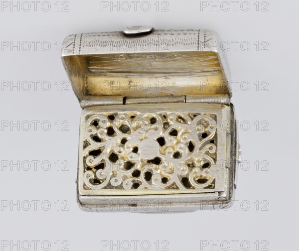 Fragrance container vinaigrette, 1834/1835. Silver vinaigrette in the form of a bag with an envelope closure and a protruding tongue; front decorated with dot-engraved flowers and leaves. Hinged lid opens to reveal an openwork insert engraved with plant scrolls. Maker: Thomas Shaw fl. 1822–ca. 1840; 1782–1845, England, Birmingham. Materials/technique: engraved silver. Function: toiletry fragrance container. Dimensions: height 2.1 cm, width 1.5 cm, depth 1 cm; weight 10 g., A small metallic rectangular box with its lid open revealing an inner compartment and a decorative pierced panel; the exterior metal appears silver-colored with engraved patterns on the lid, while the interior and the decorative panel show a golden-colored finish visible through ornate swirling cutouts; the pierced panel has a floral or scroll-like pattern with a mix of silver and gold tones, and the hinge and edges show the same silver color; the background is a plain light gray.