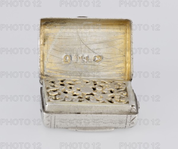 Fragrance container vinaigrette, bag-shaped with envelope closure and protruding tongue; front decorated with dot-engraved flowers and leaves; hinged lid opening to box with openwork insert decorated with plant scrolls. Silver; engraved decoration. Maker: Thomas Shaw fl. 1822–ca. 1840; inscription/marking date 1834/1835. England, Birmingham. Function: toilet accessory. Dimensions: height 2.1 cm, width 1.5 cm, depth 1.0 cm; weight 10 g., A small rectangular metal box with its lid open, showing an inner surface with a scratched texture and a golden color contrasted by silver edges; the interior of the lid has a smooth golden area with visible fine scratches. The top of the box features a decorative pierced metal insert with an ornate floral or scroll-like pattern in a mix of golden and silver tones. The exterior sides of the box are silver with subtle engraved linear patterns and slightly rounded corners. The background is a neutral light gray.