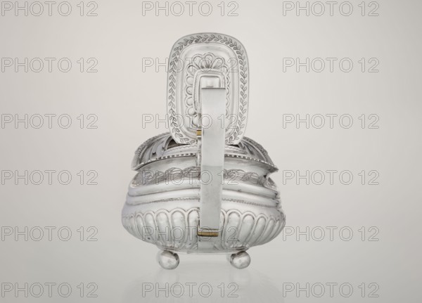 Teapot, maker: George McHattie fl. 1799–ca. 1825. English Regency style, 1819–1820, Edinburgh, Scotland. Rectangular teapot with rounded edges supported by four ball feet; decorated with hoardings ornament at the bottom, two smooth half-cylinders above, and an engraved braid band at the narrowing. Upper edge has a collar decorated with hoardings and flowers. Hinged, rectangular profiled lid edged with tendril; spout-shaped spout set high on the axis. Broken ear with bone insulators. Materials/technique: silver tea vessel. Dimensions: height 16 cm, width 28.5 cm, depth 13.5 cm; weight 735 g., A polished silver vessel with a hinged lid and a central handle sits on a reflective surface; the vessel has rounded, bulbous sides with scalloped vertical ribs and rests on a group of spherical feet, while the lid and handle feature engraved leafy and scallop patterns and decorative embossed borders in silver and a touch of gold; the background is a smooth gradient of light gray to off-white.