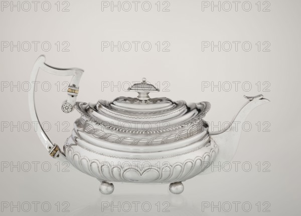 Teapot. Maker: George McHattie fl. 1799–ca. 1825, goldsmith. English Regency style, 1819–1820, Edinburgh, Scotland, Great Britain. Rectangular teapot with rounded edges, supported by four ball feet; decorated with hoardings ornament at the bottom and two smooth half-cylinders above; engraved band of braid at the narrowing; upper edge with a collar decorated with hoardings and flowers. Hinged, rectangular profiled lid edged with tendril; spout-shaped spout set high on the axis; broken ear with bone insulators. Materials/function: tea vessel. Dimensions: height 16 cm, width 28.5 cm, depth 13.5 cm, weight 735 g., A polished silver teapot with an ornate lid and a decorative finial sits on a reflective surface, showing a curved handle on one side and a long spout on the other; the body has layered, rounded ridges and embossed leaf and rope-like patterns, with scalloped and petal motifs near the base and resting on a group of rounded feet; the handle features small bands of a contrasting war...