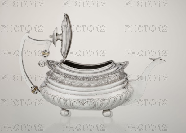Teapot, maker: George McHattie fl. 1799–ca. 1825. English Regency rectangular teapot with rounded edges, supported by four ball feet. Decorated with hoardings ornament at the bottom, above which are two smooth half-cylinders; at the narrowing an engraved band of braid. Upper edge has a collar decorated with hoardings and flowers. Hinged rectangular profiled lid edged with tendril. Spout-shaped spout set high on the axis. Broken ear with bone insulators. Tea vessel, 1819–1820, Edinburgh, Scotland. Materials/technique: goldsmith work. Dimensions: height 16 cm, width 28.5 cm, depth 13.5 cm; weight 735 g., A polished metal teapot with a hinged lid propped open sits on a reflective surface, showing a rounded body with ornate raised patterns and braided decorative bands, a curved spout ending in a small upward curl, a handle with a curved bracket and small decorative rivet elements, and three rounded ball feet; visible colors include silver, white, pale beige, and hints of muted gold on smal...