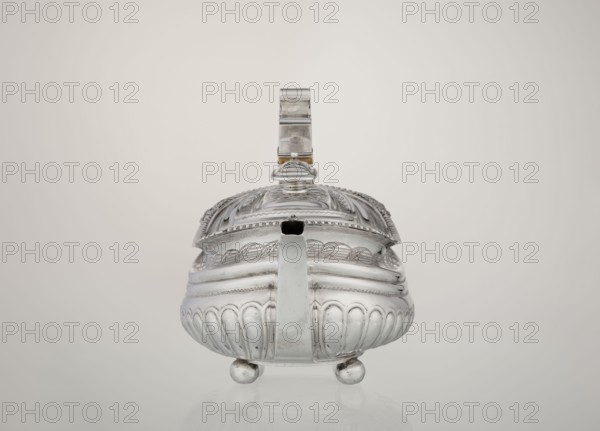 Teapot, maker: George McHattie fl. 1799–ca. 1825. English Regency rectangular teapot with rounded edges, supported by four ball feet. Decorated with hoardings ornament at the lower section, above which are two smooth half-cylinders; at the narrowing an engraved band of braid. Upper edge has a collar decorated with hoardings and flowers. Hinged, rectangular profiled lid edged with tendril; spout shaped, set high on the axis. Broken ear with bone insulators. Inscribed/date: 1819–1820, Edinburgh, Scotland, Great Britain. Material/function: tea vessel; height 16 cm, width 28.5 cm, depth 13.5 cm, weight 735 g., A shiny metallic vessel sits centered on a smooth surface against a pale beige background, featuring a rounded body with vertical fluted sections and ornate engraved patterns, a domed lid with decorative ridges and a finial on top, a broad handle rising from the rear, a short spout at the front, and three small spherical feet; visible colors include silver tones on the vessel, a hint...