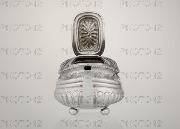Teapot, maker: George McHattie fl. 1799–ca. 1825. English Regency style, 1819–1820. Rectangular teapot with rounded edges, supported by four ball feet; decorated with hoardings ornament at the bottom and two smooth half-cylinders above; engraved braid band at the narrowing; upper edge with a collar decorated with hoardings and flowers. Hinged, rectangular profiled lid edged with tendril; high axial spout shaped spout; broken ear with bone insulators. Materials: silver tea vessel. Dimensions: height 16 cm, width 28.5 cm, depth 13.5 cm; weight 735 g. Place of manufacture: Edinburgh, Scotland, Great Britain., A metallic container with an open hinged lid sits centered against a light beige background, showing a reflective silver surface with decorative embossed patterns; the lid interior has a raised floral or fan-like motif and a small central knob, and the body features rounded fluted panels, engraved border designs, a straight handle or spout at the front, and rests on a few spherical f...