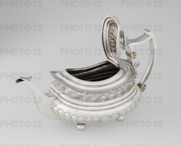 Teapot. Maker: George McHattie fl. 1799–ca. 1825, goldsmith. English Regency style, 1819–1820, Edinburgh, Scotland, Great Britain. Rectangular teapot with rounded edges, supported by four ball feet; decorated with hoardings ornament at the bottom and two smooth half-cylinders above; narrowing with an engraved band of braid; upper edge with a collar decorated with hoardings and flowers. Hinged rectangular profiled lid edged with tendril; spout-shaped spout set high on the axis; broken ear with bone insulators. Material: metal teapot/vessel. Dimensions: height 16 cm, width 28.5 cm, depth 13.5 cm; weight 735 g., A silver teapot with an open hinged lid sits against a light gray background; the teapot has a curved spout with a decorative tip, an ornate handle with small gold-colored accents, and a rounded body resting on small spherical feet; the surface shows intricate embossed and engraved patterns, scalloped and rope-like details, and a dark interior visible through the open top; visible...