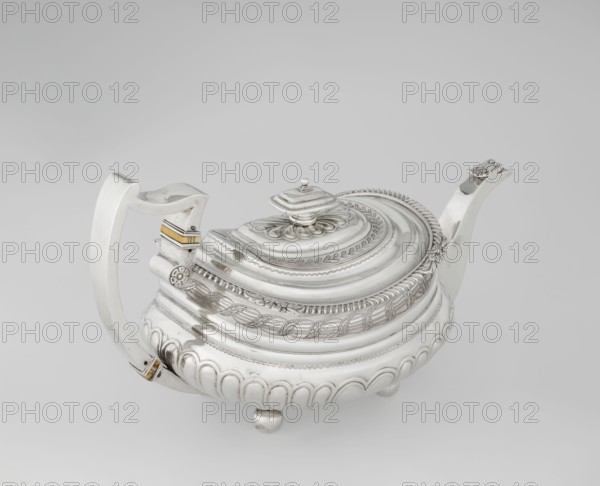 Teapot by George McHattie fl. 1799–ca. 1825, English Regency style, 1819–1820, Edinburgh, Scotland. Rectangular teapot with rounded edges supported on four ball feet. Decorated with hoardings ornament at the bottom, above which are two smooth half-cylinders; at the narrowing is an engraved band of braid. Upper edge has a collar decorated with hoardings and flowers. Hinged, rectangular profiled lid edged with tendril. Spout-shaped spout set high on the axis. Broken ear with bone insulators. Silver vessel; height 16 cm, width 28.5 cm, depth 13.5 cm, weight 735 g., A silver teapot with ornate raised patterns sits on a light grey background, showing a curved handle opposite a spout, a domed lid with a decorative finial, and a rounded body resting on small feet; the surface displays engraved bands, leaf and rope motifs, scalloped lower edges, and a small area of gold-colored accents near the handle.