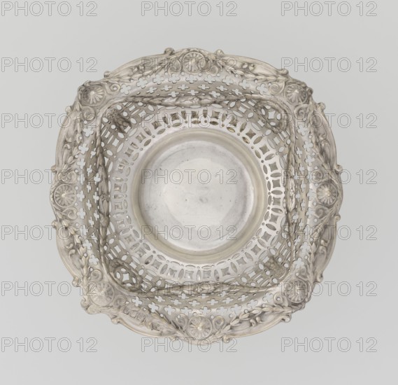 Openwork cake stand by Mappin & Webb Ltd goldsmith, plater, sellers; fl. 1859-. Openwork cake stand on a circular base with a flange, raised and decorated with relief ornament of rocaille and floral motifs; short stem supporting a circular bowl transitioning into a square one with upturned, rounded edges. Edges decorated with ovals, festoons, wreaths and rosettes. Materials/technique: openwork, relief decoration. Date: 1903/1904. Place: London, England. Object type: vessel. Dimensions: height 7.2 cm; width 14.3 cm; depth 14.3 cm; weight 112 g., A decorative metallic bowl with a round smooth center and an ornate pierced square rim, showing a group of floral and scroll motifs around the edges; the surface appears shiny and reflective with areas of subtle tarnish, and the visible colors are silver and light grey.