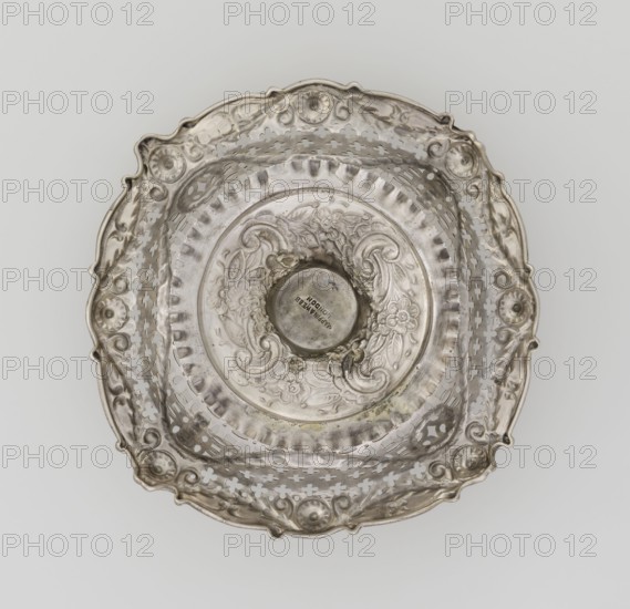 Openwork cake stand by Mappin & Webb Ltd goldsmith, plater, sellers; fl. 1859-. Openwork cake stand on a circular base with a flange, raised and decorated in relief with rocaille and floral motifs; short stem supporting a circular bowl transitioning into a square one with upturned, rounded edges. Edges decorated with ovals, festoons, wreaths and rosettes. Materials/technique: openwork, relief decorations. Date: 1903/1904. Place: London, England, Great Britain. Function: vessel. Dimensions: height 7.2 cm; width 14.3 cm; depth 14.3 cm; weight 112 g., A decorative metal bowl viewed from above with an ornate scalloped rim and pierced sides, showing floral and scrollwork patterns in relief; the metal surface appears reflective and aged with areas of tarnish in shades of silvery gray and darker gray, with subtle hints of warm beige where the metal shows wear; the bowl has a central circular depression and concentric decorative bands with embossed flowers and curved motifs, and the background...