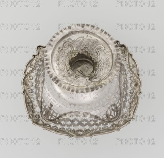 Openwork cake stand, Mappin & Webb Ltd goldsmith, plater, sellers; fl. 1859- author. Openwork cake stand on a circular base with a flange, raised and decorated with a relief ornament with rocaille and floral motifs, with a short stem supporting a circular bowl transitioning into a square one with upturned, rounded edges; edges decorated with ovals, festoons, wreaths and rosettes. Materials/technique: openwork, relief decorations. Style/ornament: rosette, geometric ornaments, festoons, rocaille. Date: 1903/1904. Place: London, England Great Britain. Function: vessel. Dimensions: height 7.2 cm; width 14.3 cm; depth 14.3 cm; weight 112 g., A silver-colored ornate metal object viewed from above, featuring a round central well with embossed floral and scrollwork patterns and a scalloped rim, surrounded by a square tray-like base with pierced lattice and cross-shaped cutouts, decorative raised edges with curved outlines and floral motifs, and areas of tarnish and subtle darker gray patina ag...