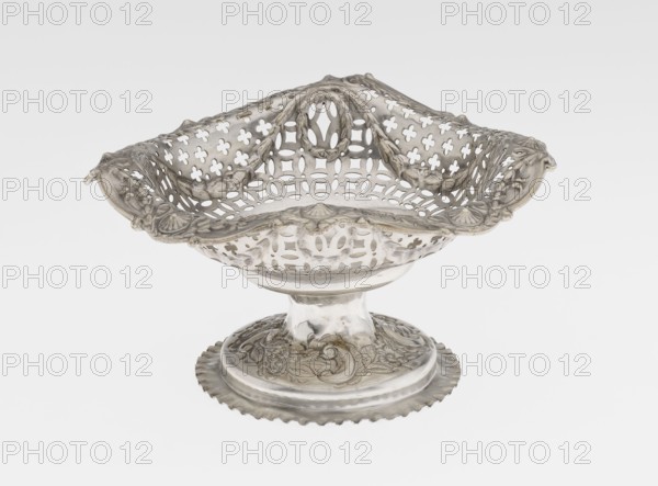 Openwork cake stand by Mappin & Webb Ltd goldsmith, plater, sellers; fl. 1859-. Openwork cake stand on a circular base with a flange, raised and decorated in relief with rocaille and floral motifs. Short stem supports a circular bowl transitioning into a square one with upturned, rounded edges, decorated with ovals, festoons, wreaths and rosettes. Materials/technique: openwork, relief decoration. Date: 1903/1904. Place: London, England. Object type: vessel. Dimensions: height 7.2 cm; width 14.3 cm; depth 14.3 cm; weight 112 g., A decorative metal bowl with an ornate pierced lattice design and scalloped rim sits on a round pedestal base; the bowl and base show detailed embossed floral and scroll motifs and a draped, wavy edge. The object appears in metallic silver tones with areas of darker patina and subtle reflections, set against a plain light gray to white background.