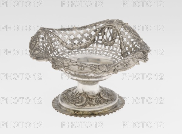 Openwork cake stand by Mappin & Webb Ltd goldsmith, plater, sellers; fl. 1859-. Openwork cake stand on a circular base with a flange, raised and decorated with a relief ornament featuring rocaille and floral motifs; a short stem supports a circular bowl transitioning into a square one with upturned, rounded edges decorated with ovals, festoons, wreaths and rosettes. Materials/technique: openwork, relief decorations. Date: 1903/1904. Place: London, England, Great Britain. Function: vessel. Dimensions: height 7.2 cm; width 14.3 cm; depth 14.3 cm; weight 112 g., A decorative metal bowl with an ornate openwork rim sits on a pedestal base; the bowl has intricate pierced patterns and carved floral and scroll motifs around its edge and on the base, with a scalloped outer rim and draped ornamental details visible along the upper rim. The surface shows a metallic sheen with areas that appear slightly darker or tarnished, displaying mainly silver tones with hints of gray.