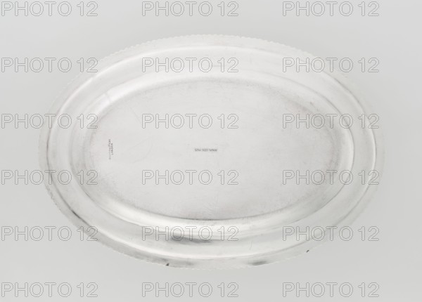 Oval platter by Mappin & Webb Ltd goldsmith, silversmith, dealer, 1904/1905. Oval platter with smooth mirror, slightly raised collar surrounded by a godron half-cylinder; on the collar a four-field coat of arms under an earl's coronet. Coats of arms, godron ornaments, rank crowns heraldry. England; Sheffield, Great Britain. Dish. Height 2 cm; width 33 cm; depth 22.5 cm; weight 625 g., A large oval metal tray fills the frame, showing a smooth, slightly worn surface with a raised rim and scalloped outer edge; the tray appears silvery and metallic with areas of subtle gray shading and faint darker scuffs and scratches, and the background is a light neutral gray.